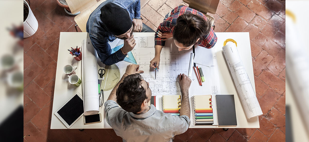 Building Designer discussing details of proposed home or renovation with two clients. Rolls of plans, pens, and set squares are visible on the table