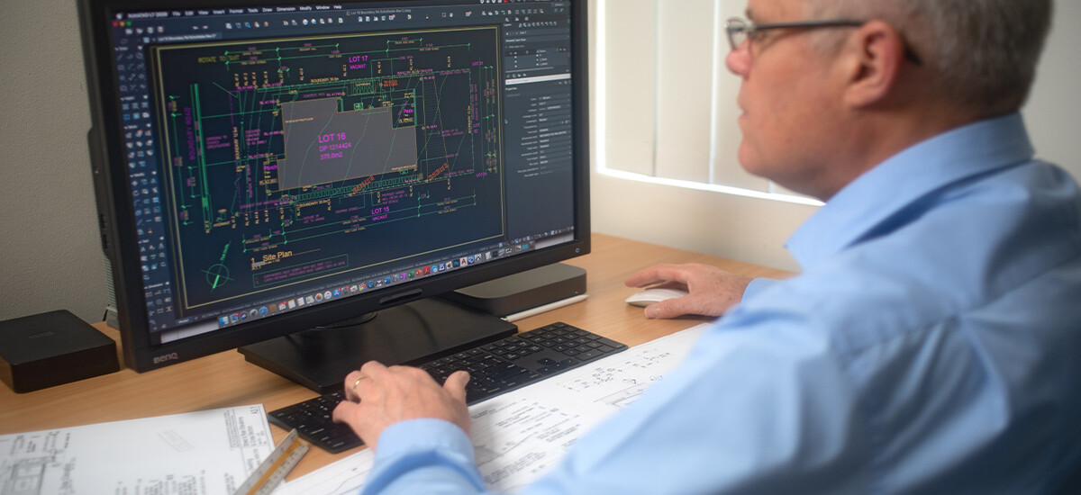 Architectural Draftsman at Work Sitting at Desk Using Computer Aided Drafting.  Site Plan visible on large screen, with printed plans and scale rule on desk.