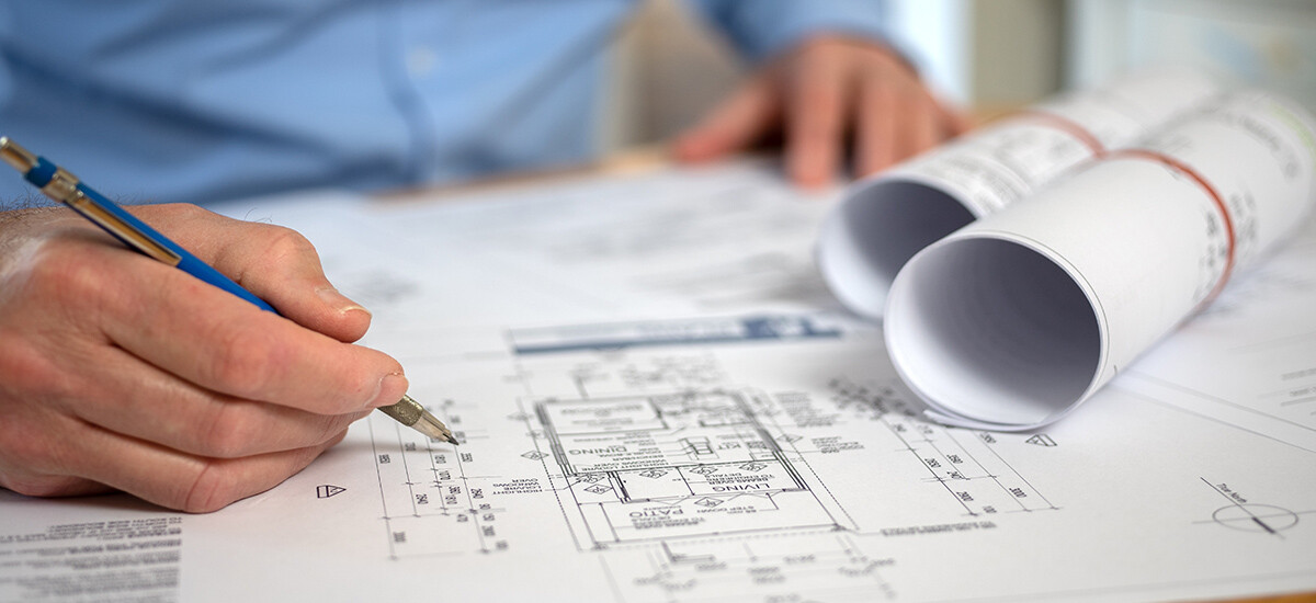 Carefully Checking Architectural Plans in Office. The Building Designer, pencil in hand, carefully checking and marking up architectural floor plan in office, with rolled plans on desk.